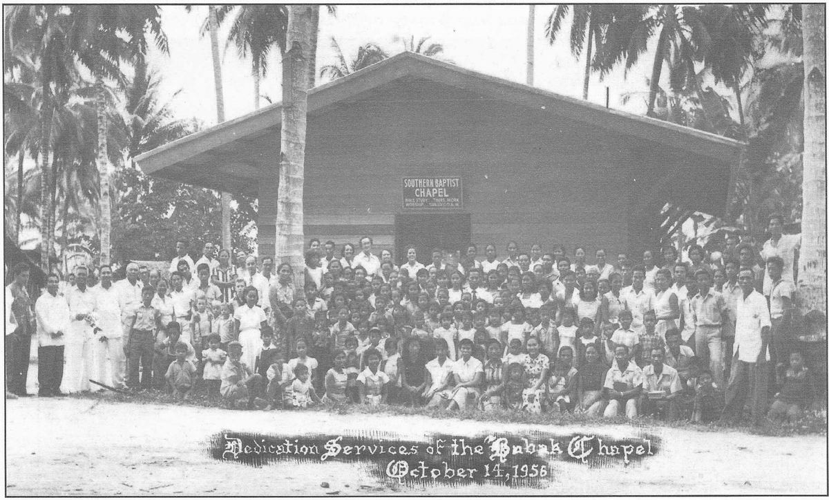 Crowd assembled in front of a newly built wooden chapel among palm trees for its dedication.